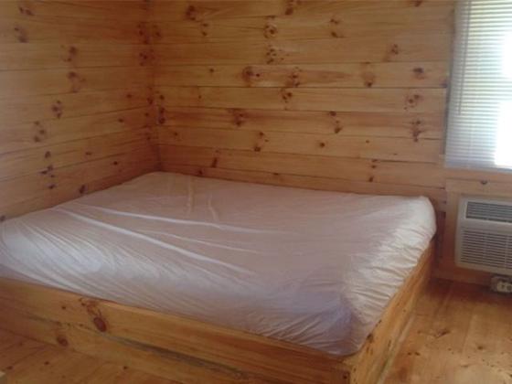 Bed with a white mattress cover in a pine-paneled corner, with a wall air conditioner