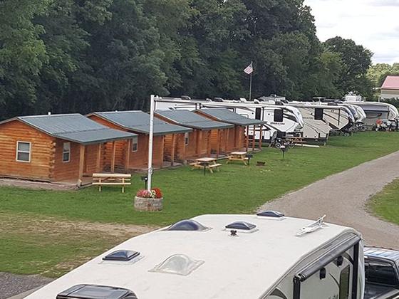 Row of small log cabins with picnic tables and a flagpole, beside parked RVs