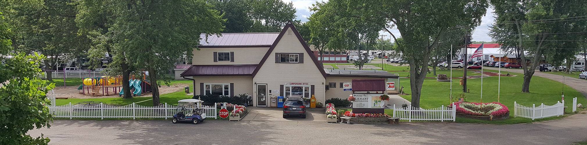 The Elkhart Campground office and store behind a white picket fence, with flower beds, a playground, and shade trees